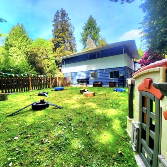 The fenced back garden with lawn, tall trees, a playhouse and tree-stump seats